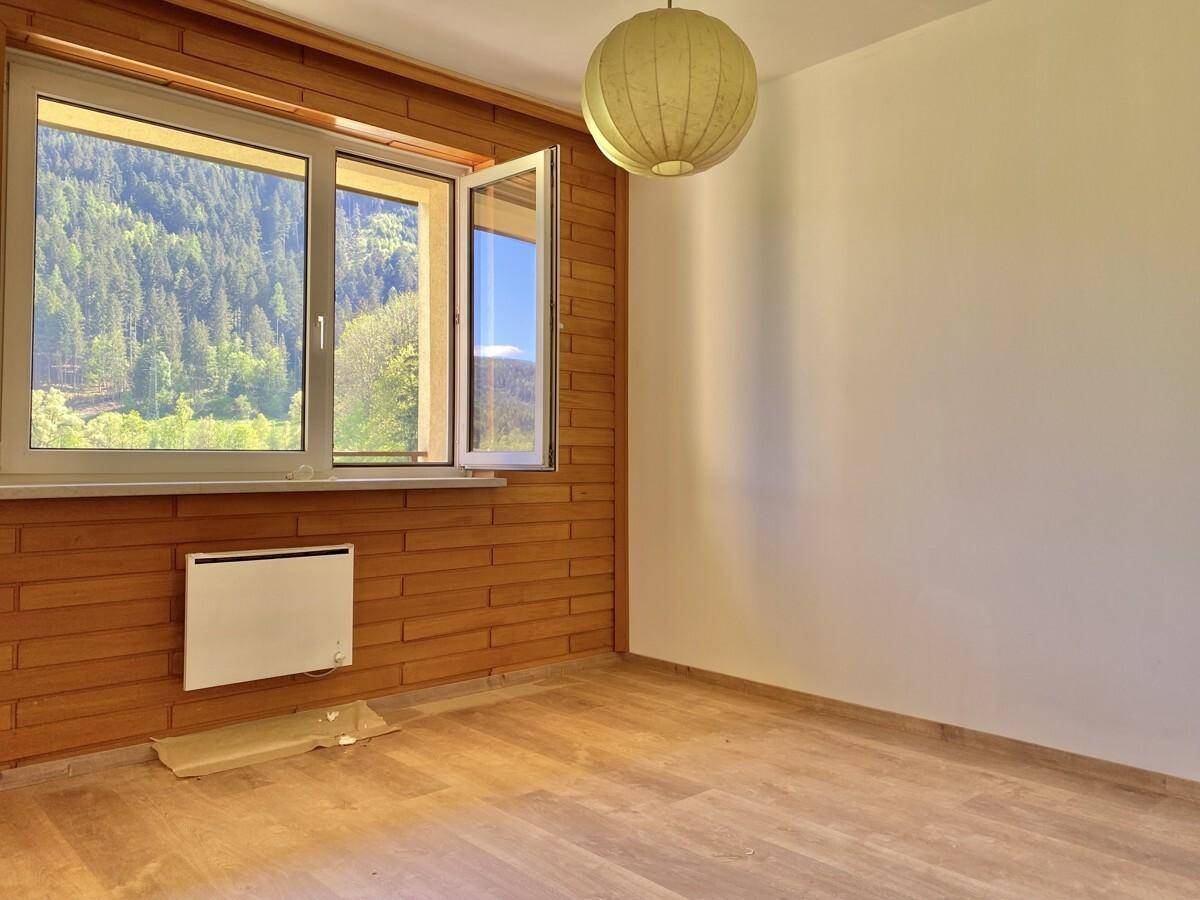 Helles Zimmer mit Holzwand, großem Fenster und Blick auf die bewaldeten Berge.