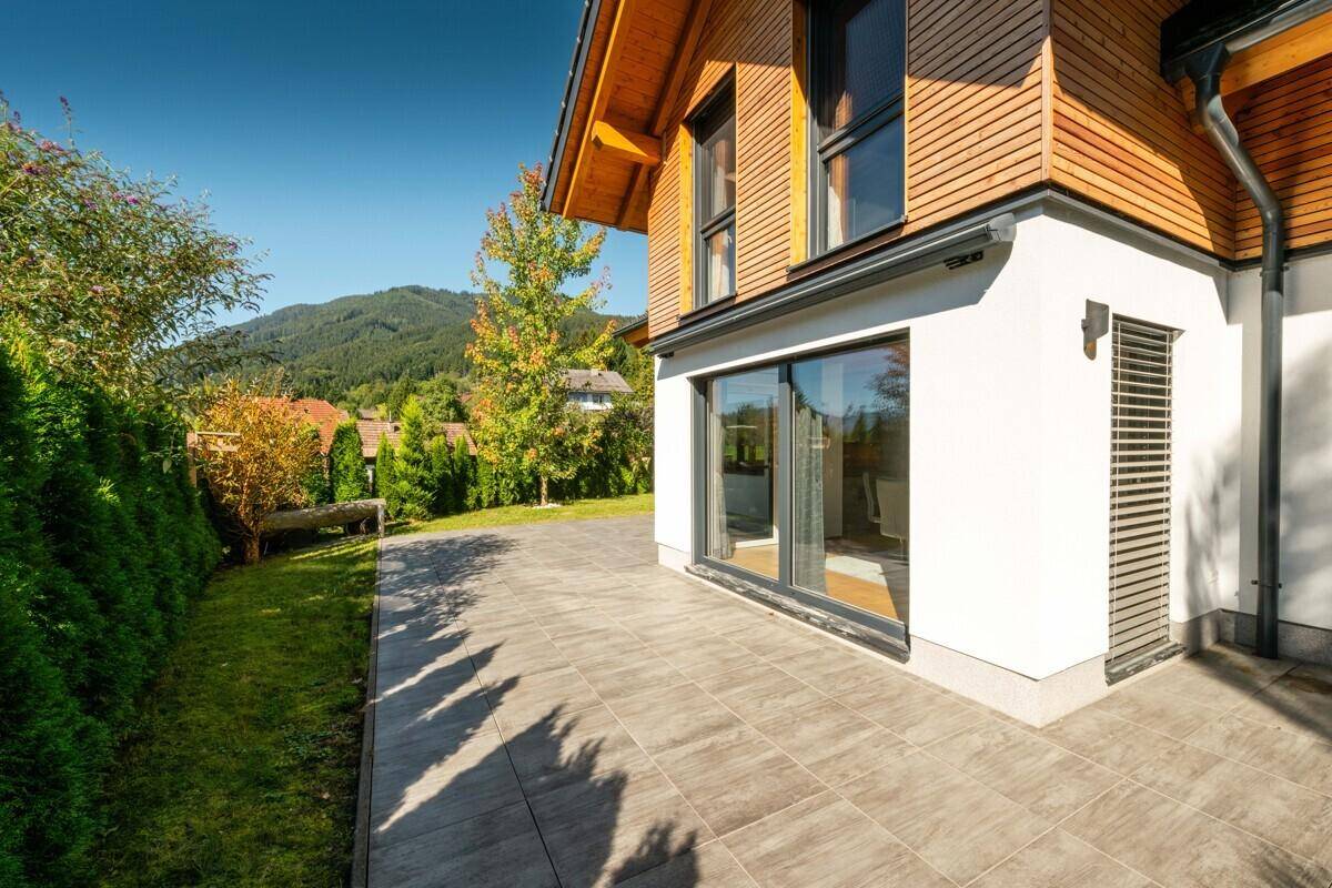 Lange, gepflasterte Terrasse entlang des Hauses mit großen Schiebetüren und einer grünen Hecke.