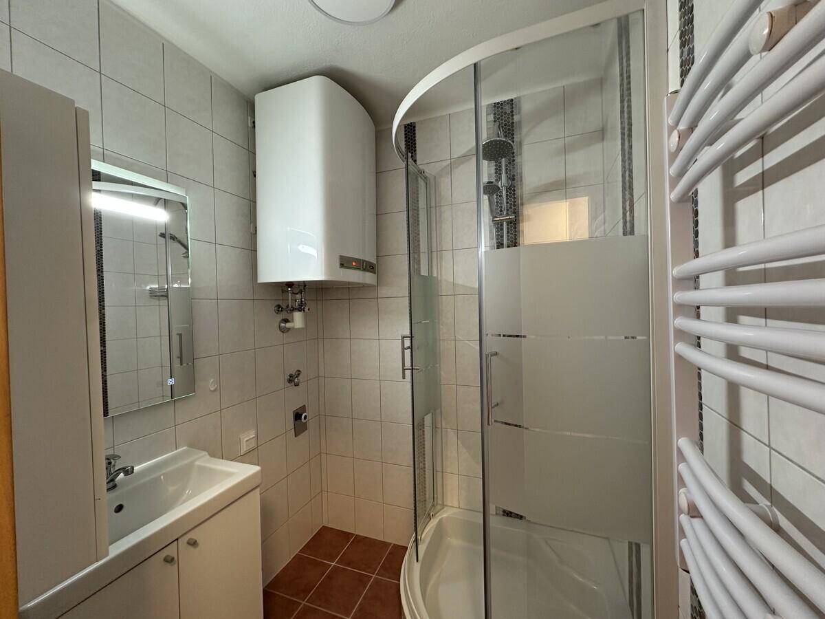 Kleines Badezimmer mit Duschkabine, Waschbecken und Wandheizkörper, komplett gefliest.