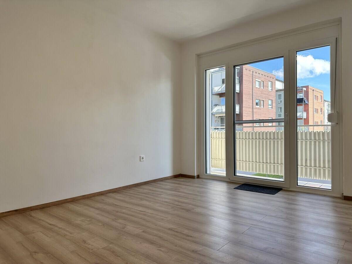 Leerer Raum mit Laminatboden und Zugang zu einem Balkon mit Blick auf andere Gebäude.