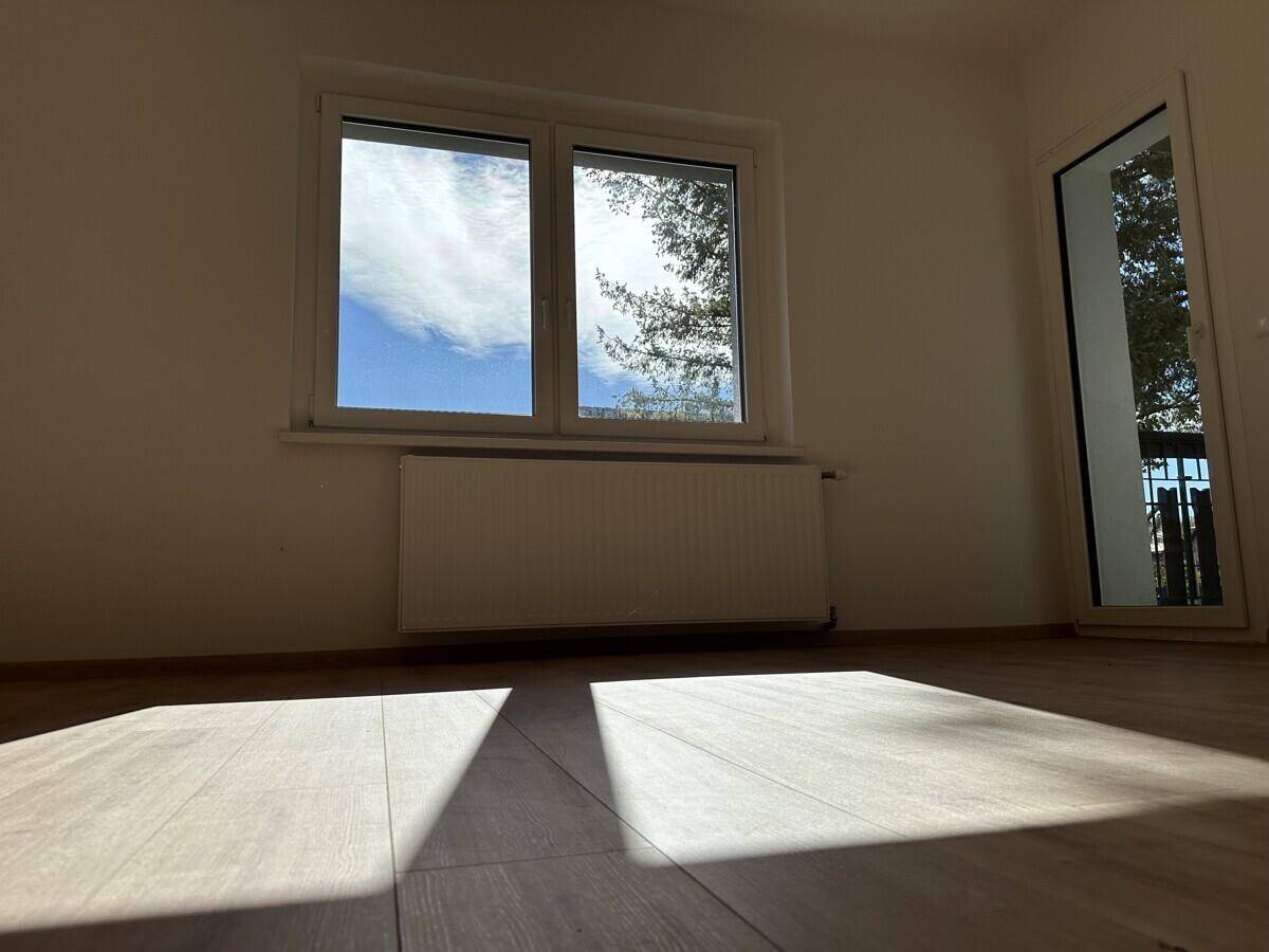 Helles Zimmer mit Holzboden, großem Fenster und Zugang zu einem Balkon.