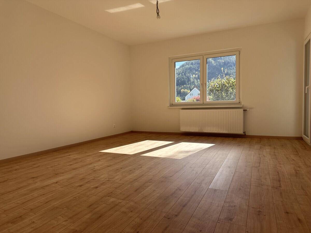 Geräumiges, leeres Zimmer mit Holzboden und Blick auf grüne Hügel durch das Fenster.