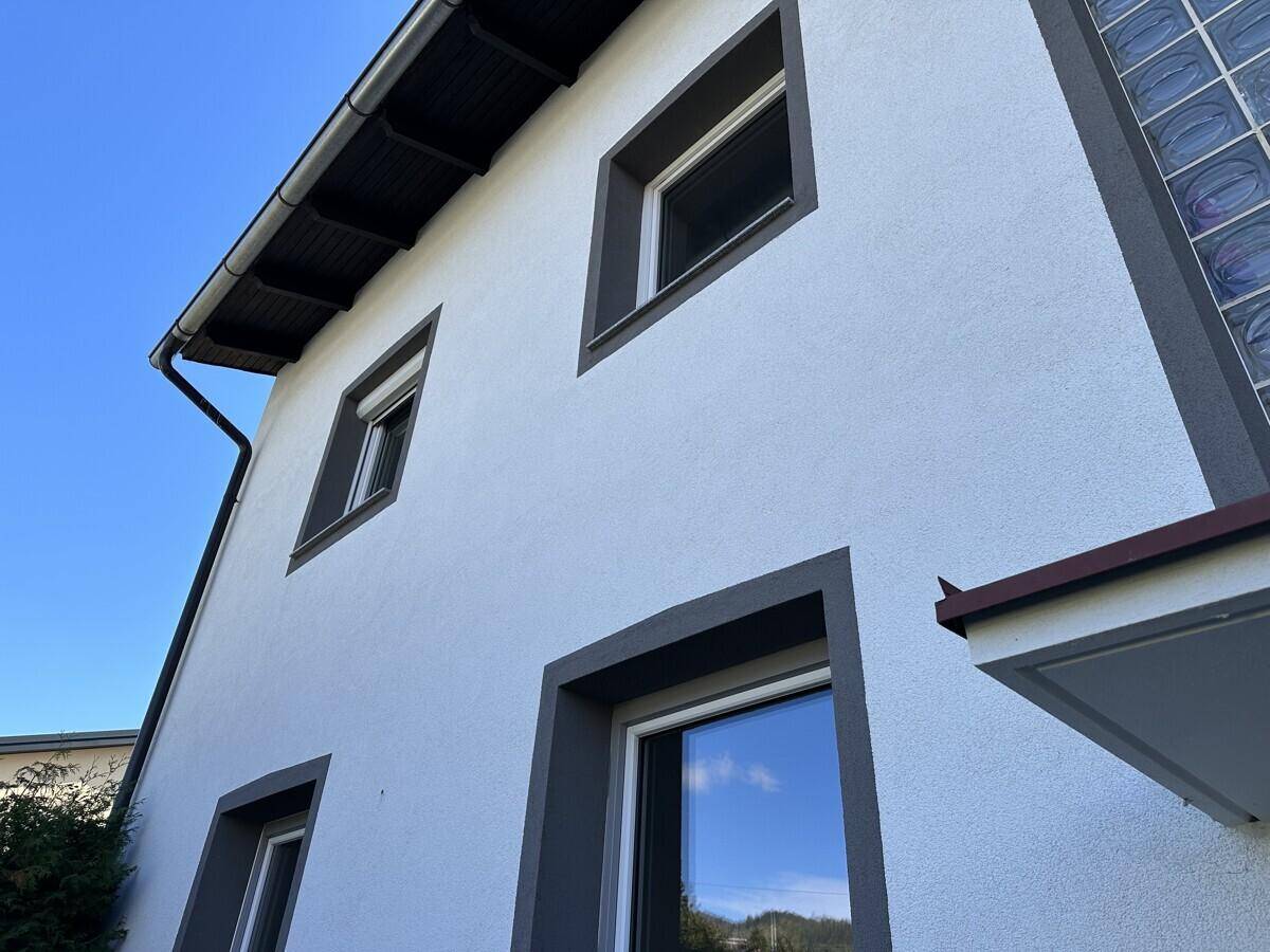 Fassade eines Wohnhauses mit weißen Wänden, grauen Fensterrahmen und Dachrinne.
