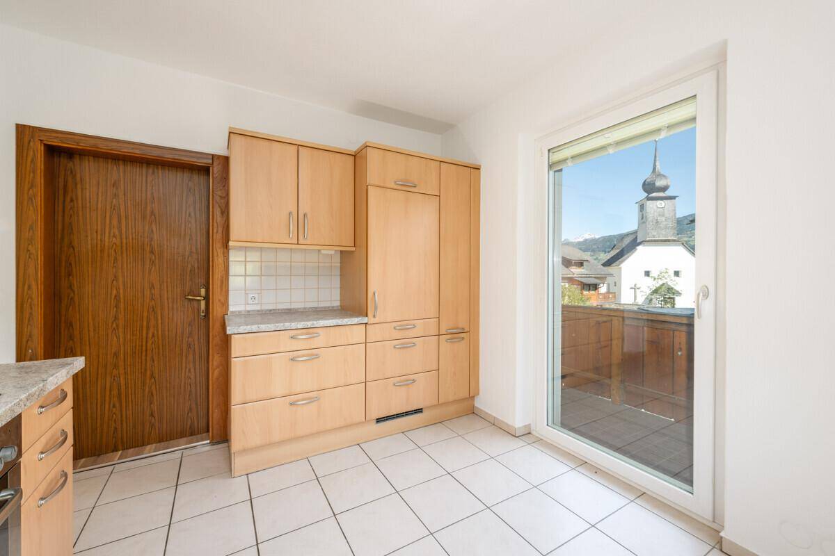 Küche mit hellen Holzschränken, Fliesenboden und Zugang zu einem Balkon mit Blick auf die Kirche.