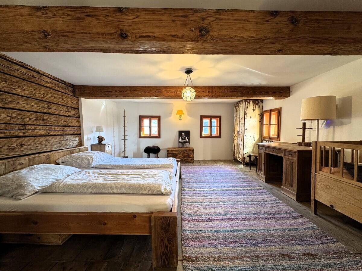 Helles Schlafzimmer mit Holzbett, Schreibtisch und farbenfrohem Teppich, unter rustikalen Deckenbalken.