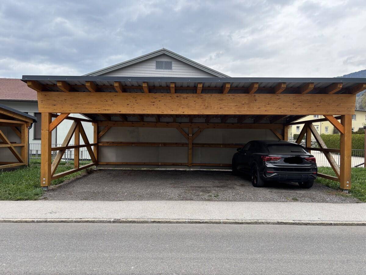 Holzcarport mit einem schwarzen Auto darunter, auf einem Schotterparkplatz.