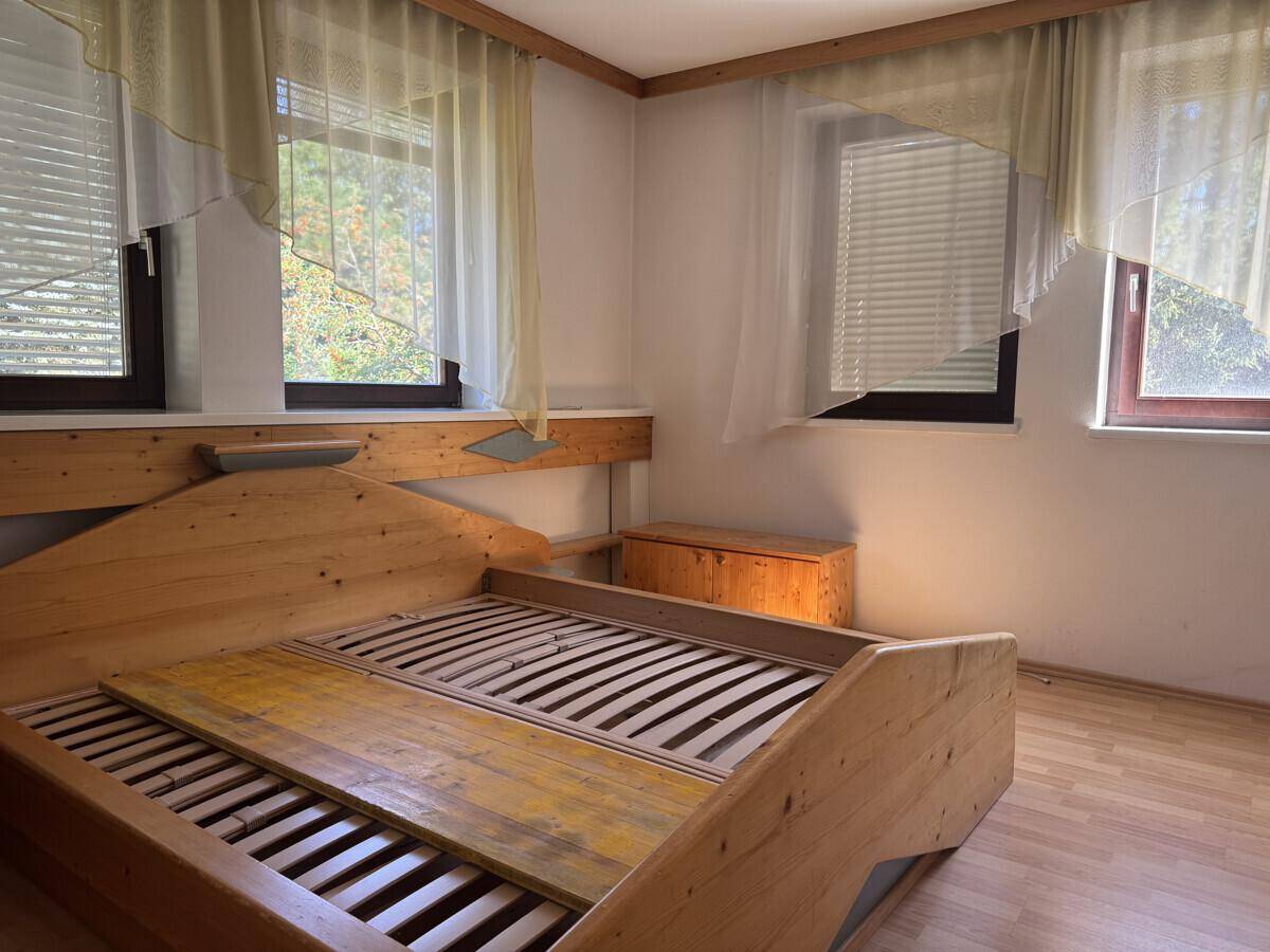 Schlafzimmer mit Holzbettrahmen, mehreren Fenstern und hellen Vorhängen.