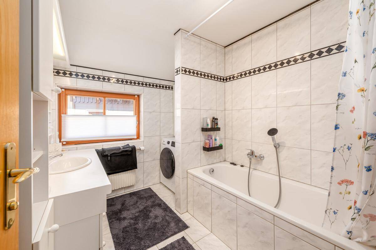 Badezimmer mit Badewanne, Einzelwaschbecken, Waschmaschine und gefliesten Wänden.