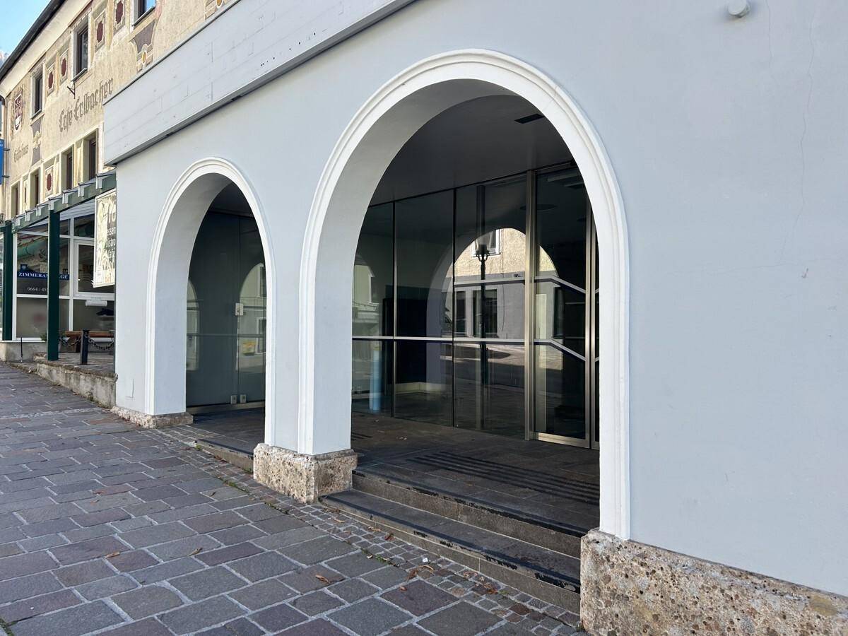 Eingangsbereich eines Gebäudes mit markanten Bögen und Glasfassade, davor ein gepflasterter Gehweg.