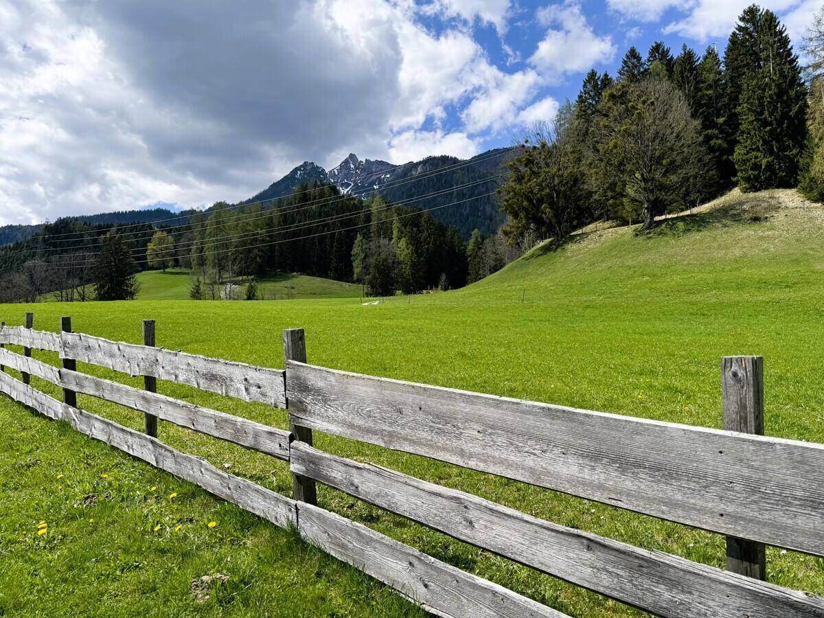 Rustikaler Holzzaun im Vordergrund mit grüner Landschaft und Bergen unter blauem Himmel.
