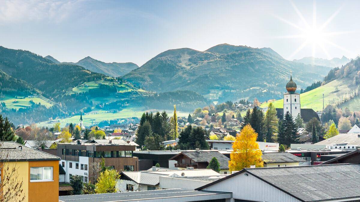 Sonniger Blick auf ein charmantes Dorf mit Kirche, umgeben von sanften Hügeln und Bergen.