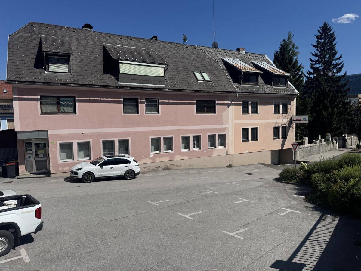 Gebäudeansicht mit Parkplatz, einem weißen SUV und der umliegenden Berglandschaft.
