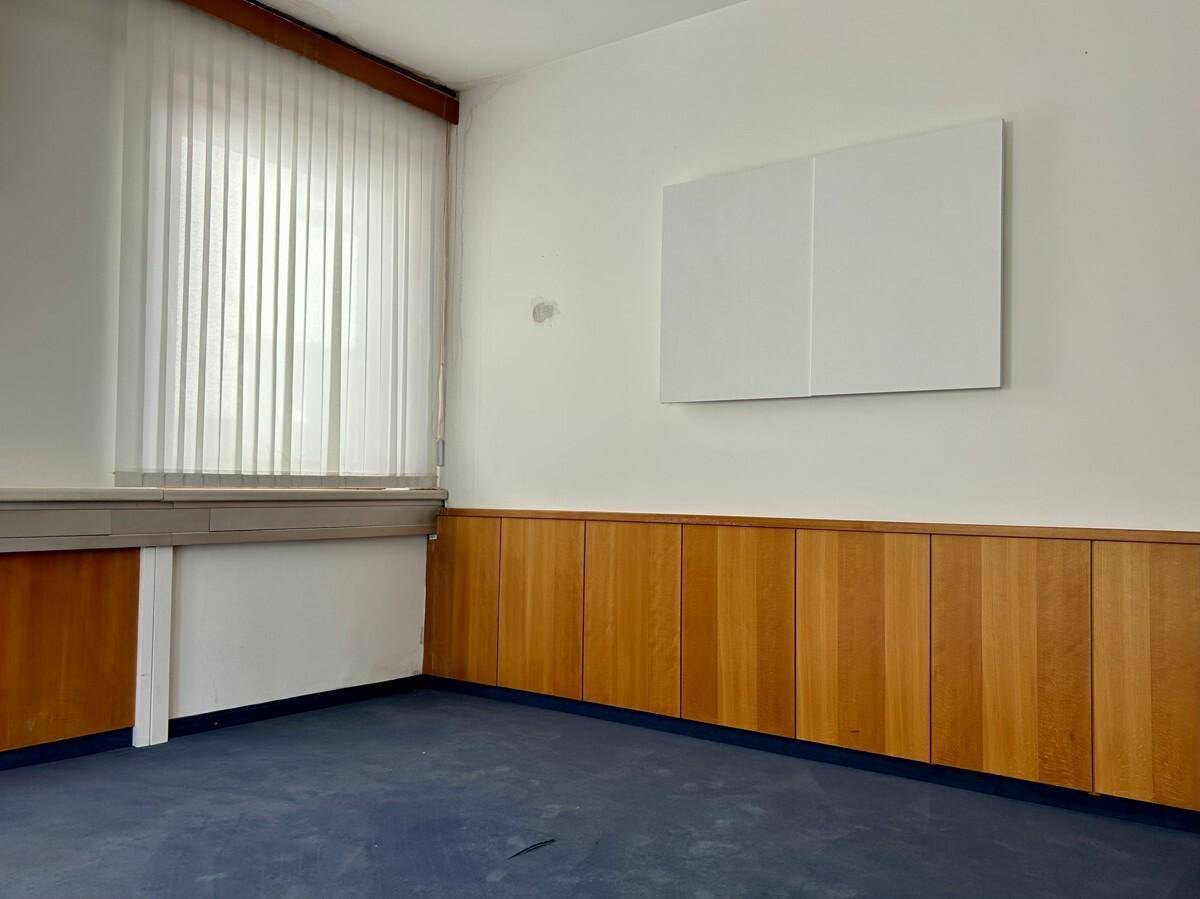 Büroraum mit blauem Teppichboden, Fenster mit Jalousien und Holzvertäfelung an der Wand.
