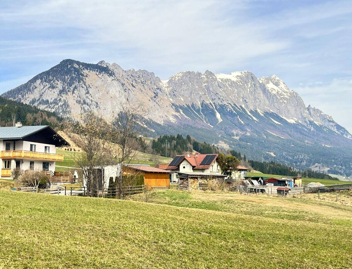 Idyllische Landschaft mit Wohnhäusern, grünen Wiesen und einer imposanten Bergkette unter blauem Himmel.