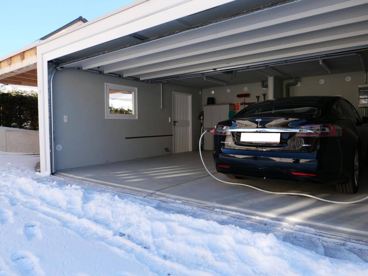 Offene Garage mit einem ladenden Elektrofahrzeug und schneebedeckter Zufahrt im Winter.