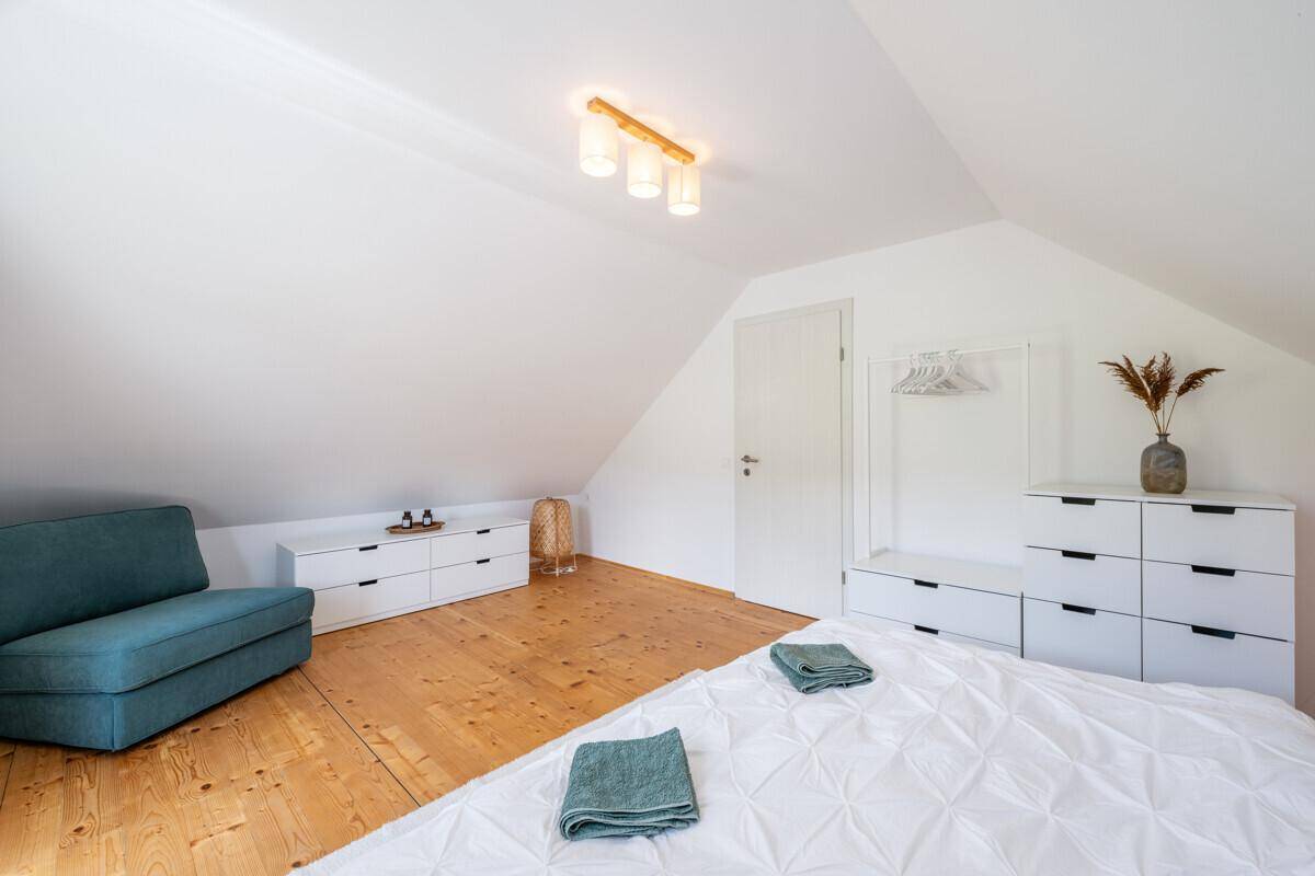 Helles Schlafzimmer im Dachgeschoss mit Holzboden, weißem Bett und modernem Sessel.