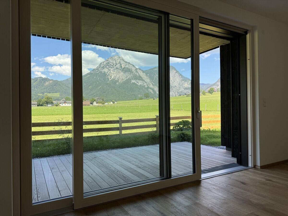 Heller Wohnbereich mit großen Schiebetüren, die auf eine Holzterrasse und zu den Bergen führen.