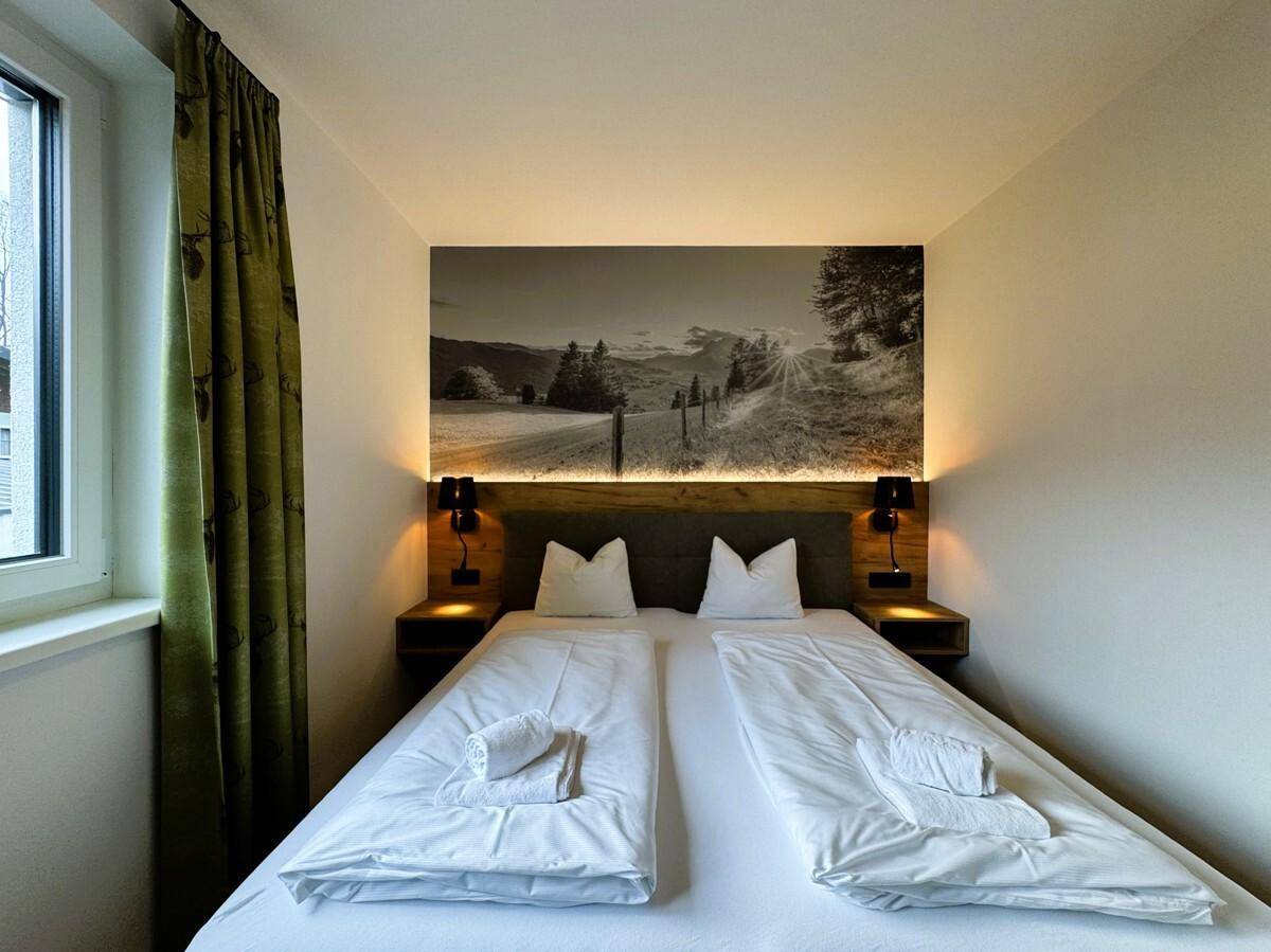 Schlafzimmer mit Doppelbett, Holzboden und Wandbild einer sonnigen Berglandschaft.
