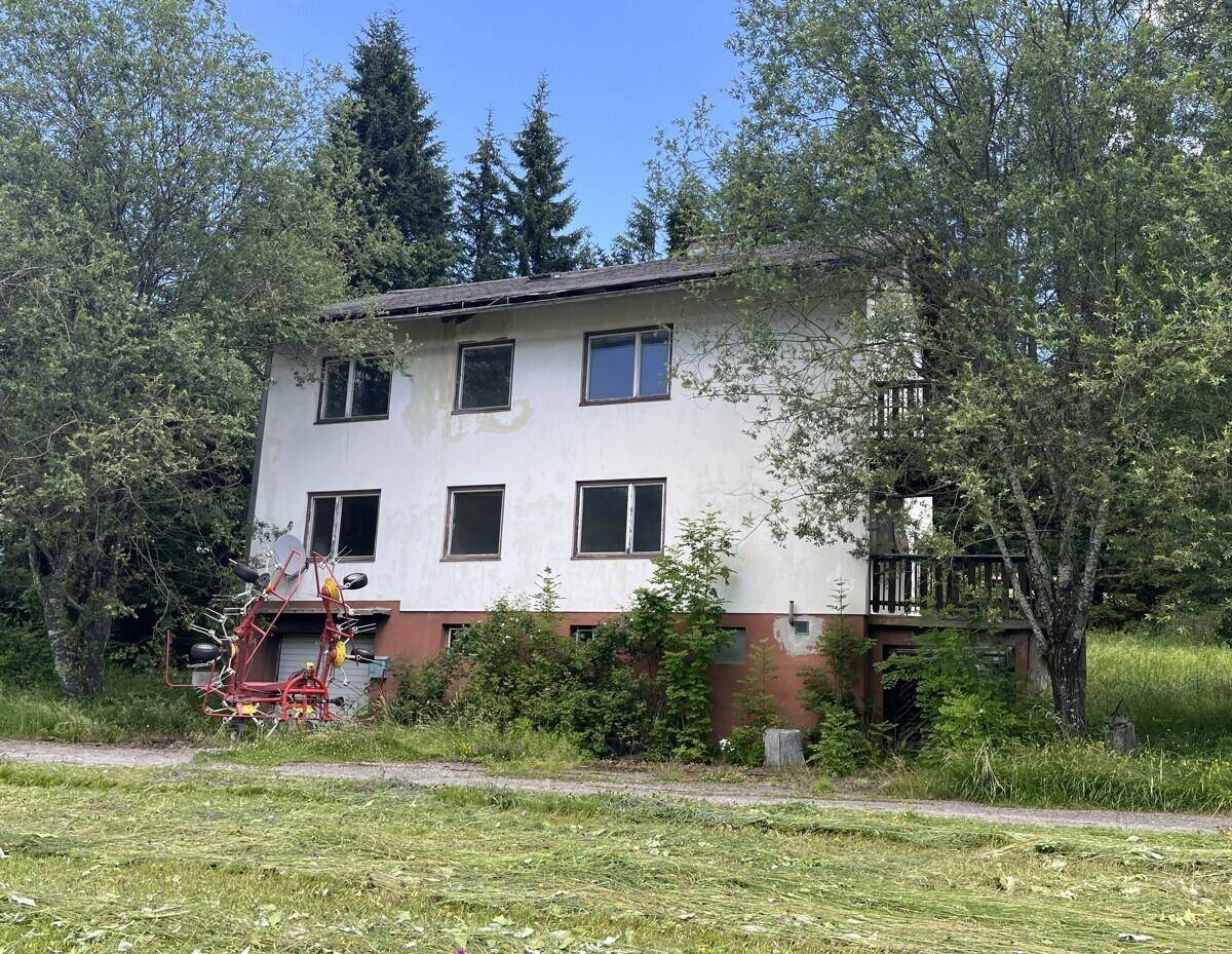 Rückseite des Wohnhauses mit Bäumen, grüner Wiese und landwirtschaftlichem Gerät im Vordergrund.