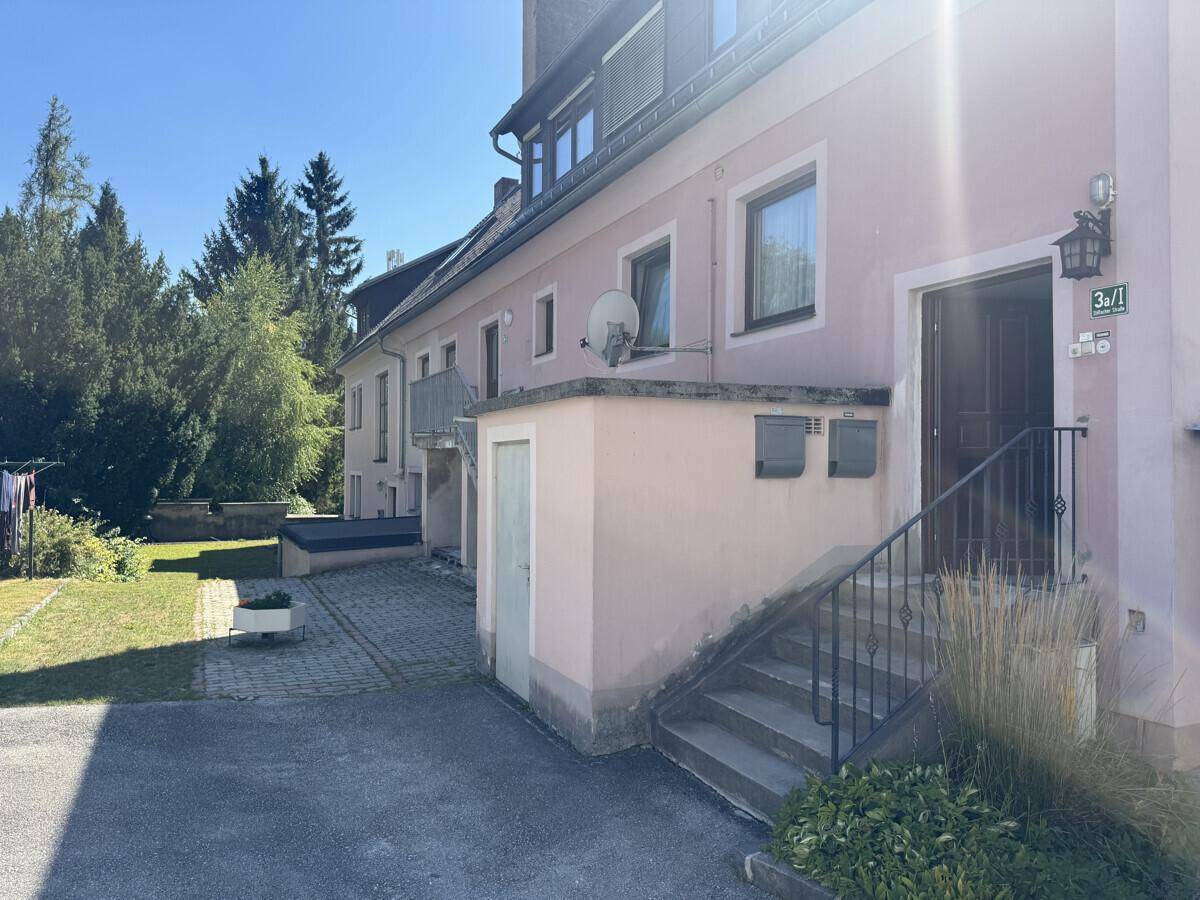 Seitlicher Eingang des Gebäudes mit Treppe, Briefkästen und einem kleinen Gartenbereich.