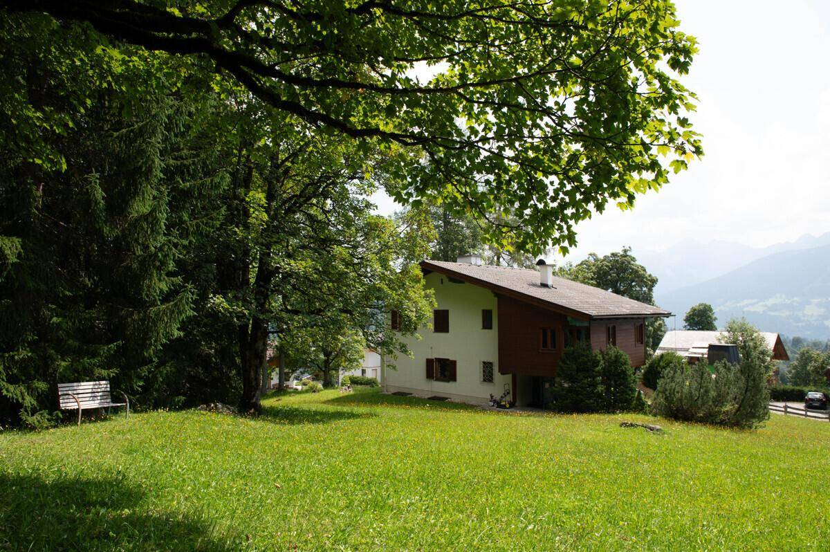 Haus mit großem Rasen, alten Bäumen und einer Bank, umgeben von grüner Natur und Bergblick.