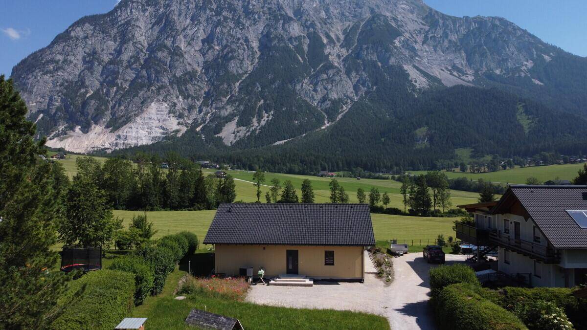 Luftaufnahme eines Hauses mit dunklem Dach, eingebettet in grüne Felder und majestätische Berge.
