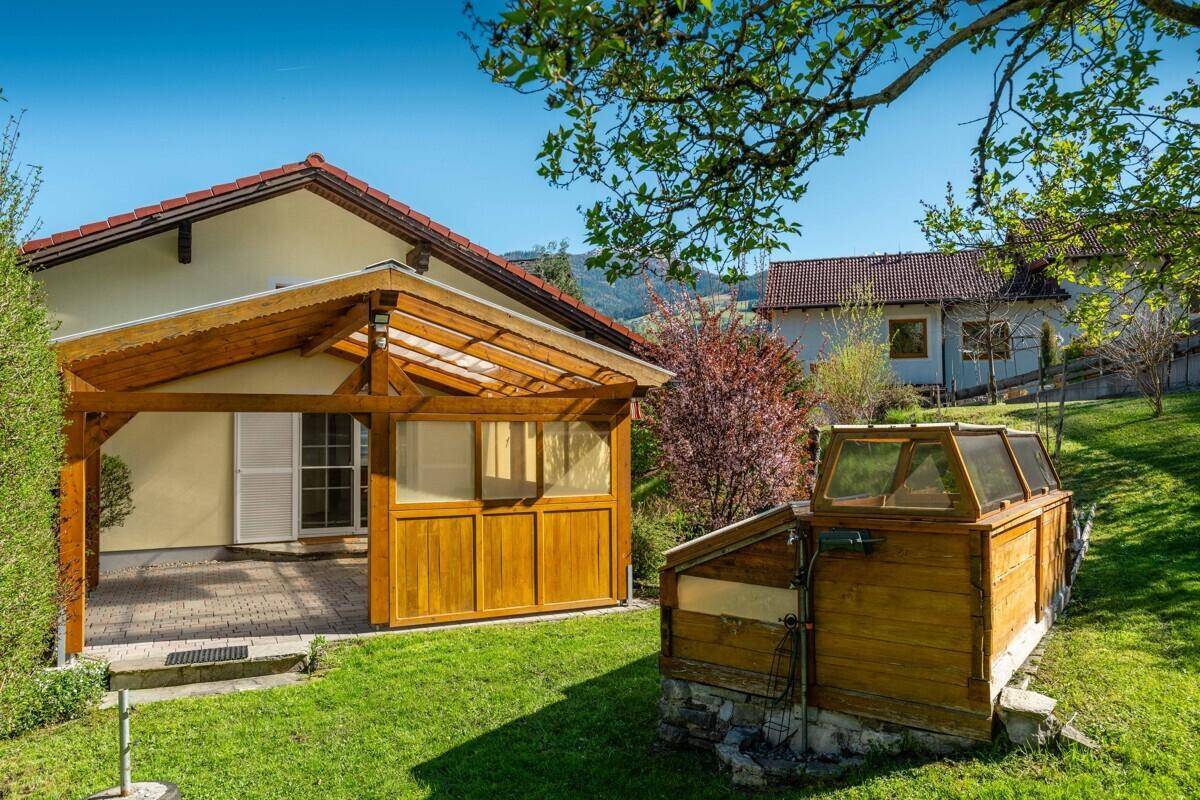 Grüner Gartenbereich mit überdachter Terrasse und einem kleinen Gewächshaus, sonnig.