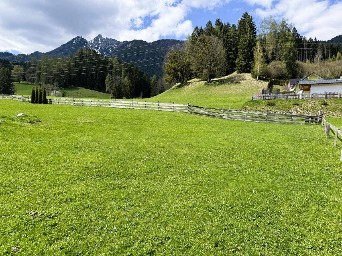 Weitläufige grüne Wiesenfläche mit Holzzaun und bewaldeten Bergen im Hintergrund unter blauem Himmel.