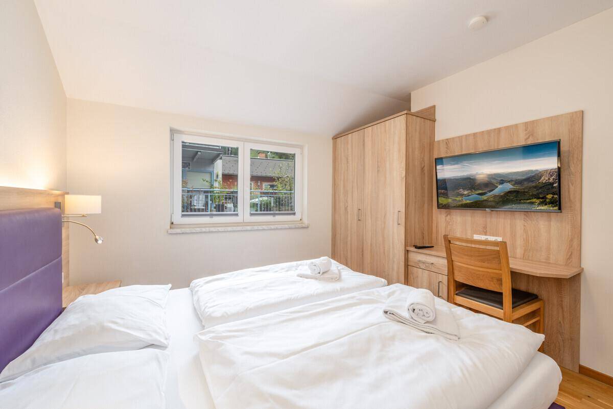 Helles Schlafzimmer mit Doppelbett, Holzmöbeln und einem an der Wand montierten Fernseher.