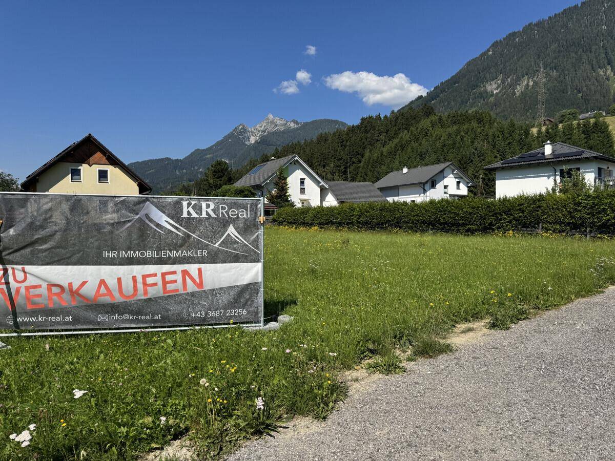 Verkaufsschild auf einem grünen Grundstück mit Wohnhäusern und Bergen im Hintergrund.