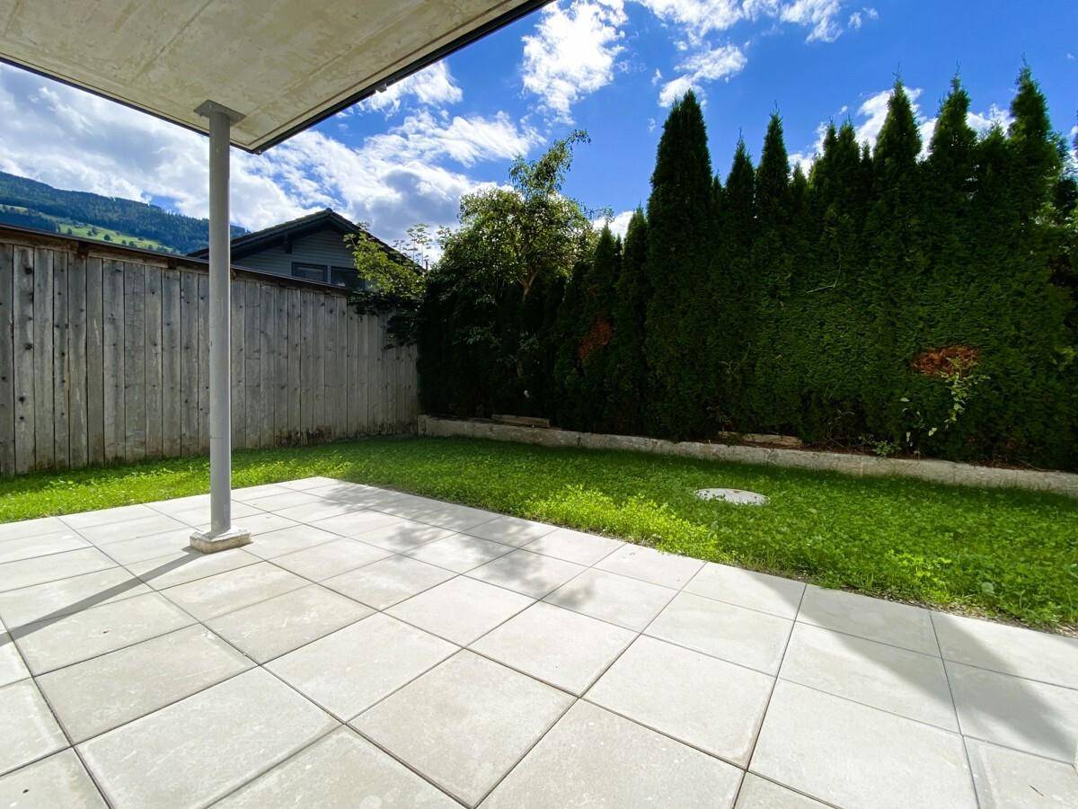Gepflasterte Terrasse mit Rasenfläche und hohen Hecken, umgeben von grüner Natur.