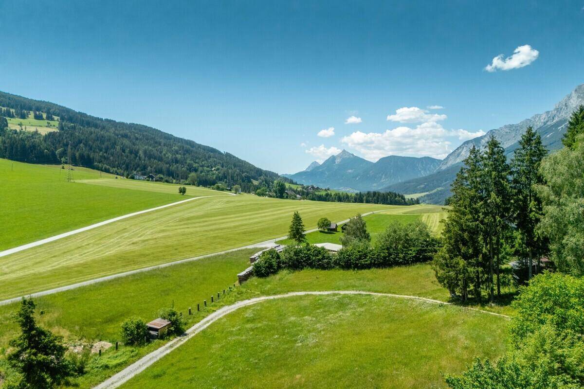 Weitläufige grüne Landschaft mit sanften Hügeln, Feldern und Bergen am Horizont unter blauem Himmel.