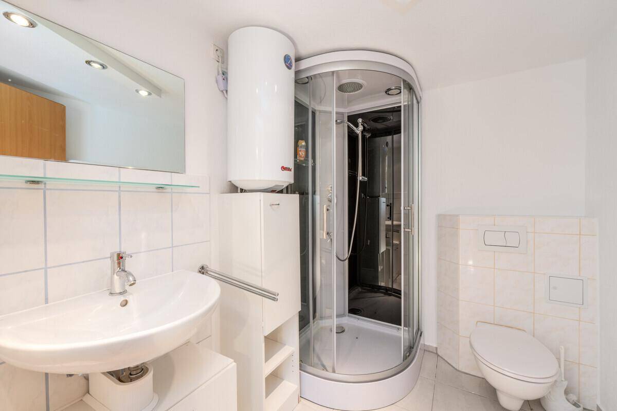 Kleines Badezimmer mit Duschkabine, Waschbecken und Toilette, mit Warmwasserboiler.