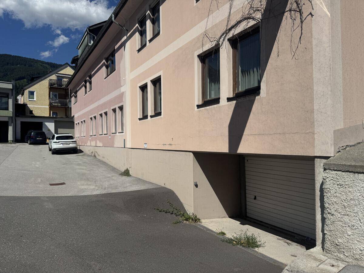 Seitenansicht des Gebäudes mit Garageneinfahrt, Parkplatz und bergiger Umgebung.