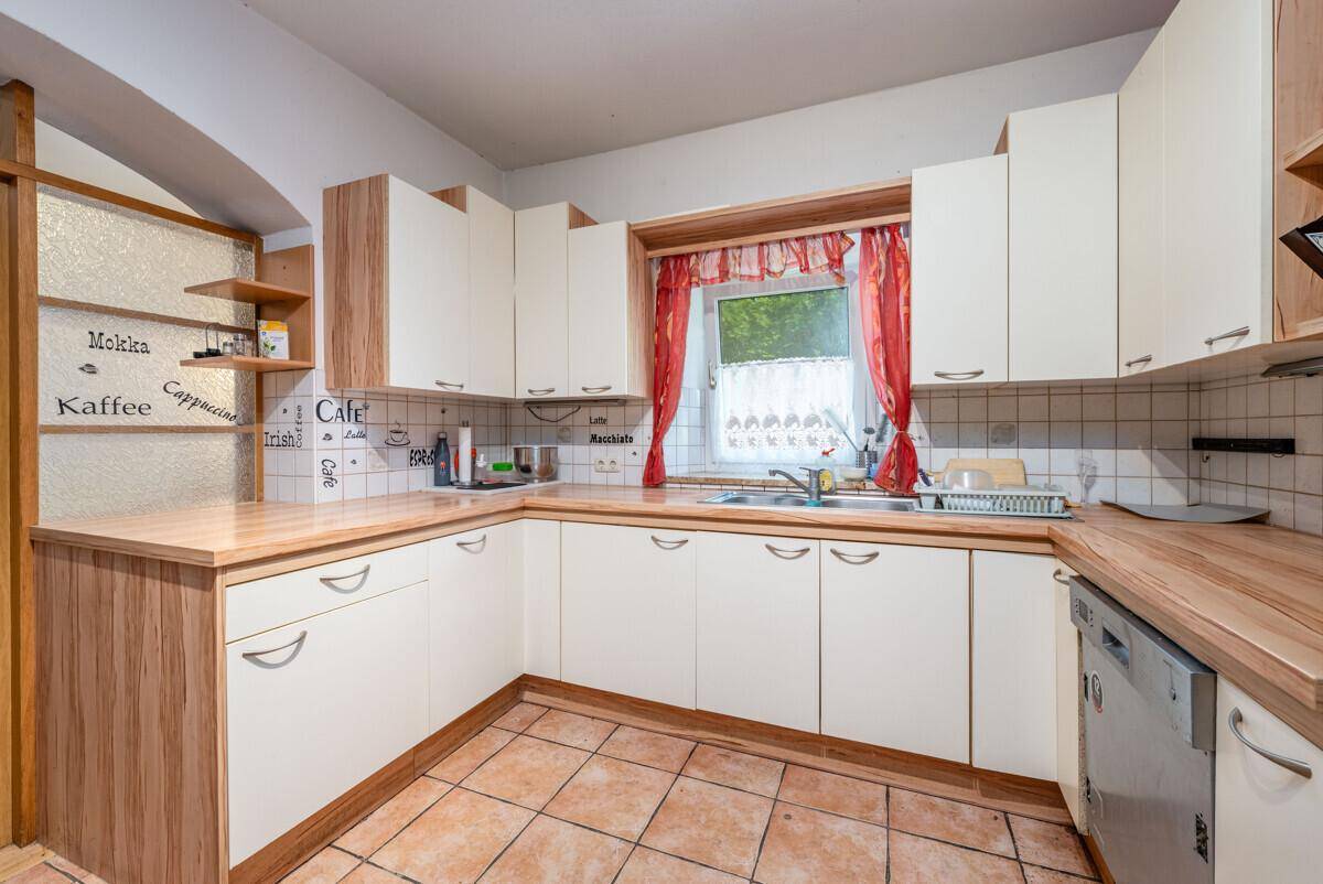 Winkelküche mit hellen Holzfronten, gefliestem Boden und Fenster über der Spüle, praktisch eingerichtet.
