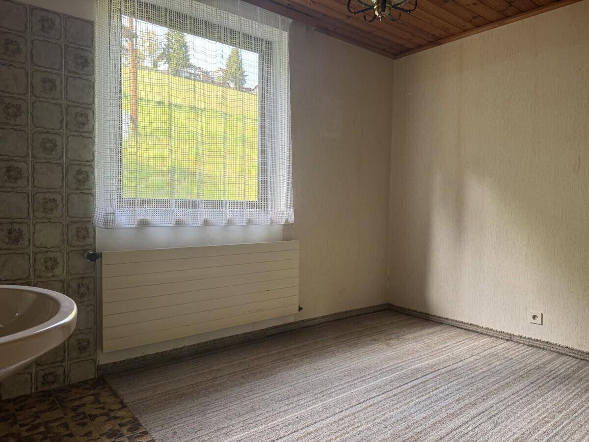 Zimmer mit Teppichboden, Holzdecke, Fenster und einem Waschbecken, in einfachem Zustand.