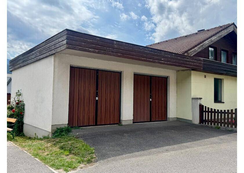 Doppelgarage mit zwei Holztoren und weißer Fassade, neben einem Wohnhaus.