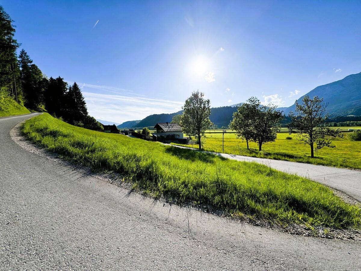 Asphaltierte Straße schlängelt sich durch eine sonnige, grüne Landschaft mit Häusern und Bergen im Hintergrund.