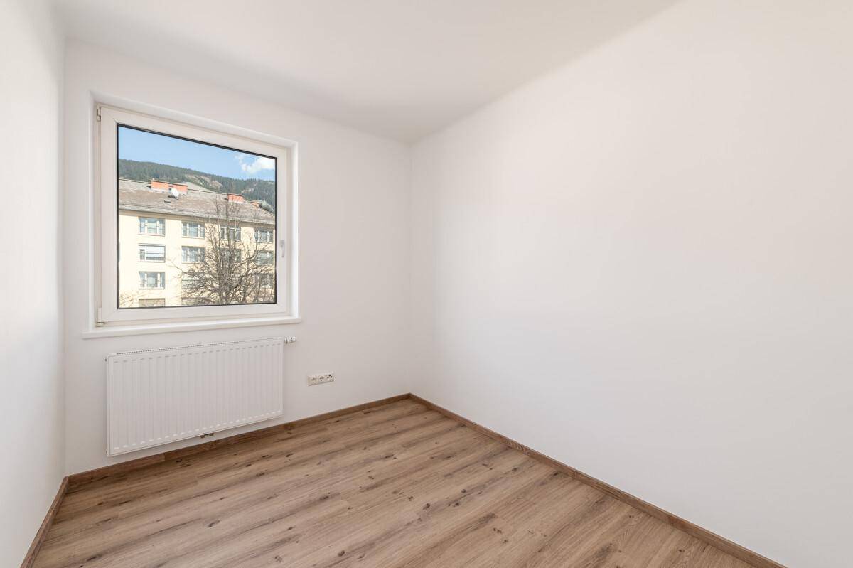 Gemütliches Zimmer mit Holzboden, weißen Wänden und einem Fenster mit Blick ins Grüne.