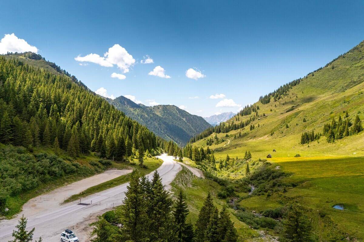 Malerische Aussicht auf eine kurvenreiche Straße durch ein grünes Bergtal unter blauem Himmel.