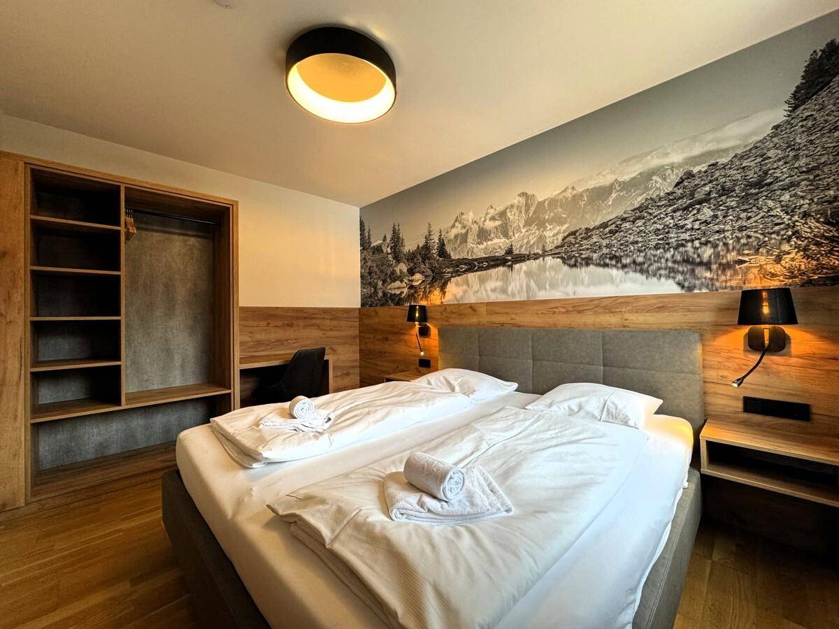 Schlafzimmer mit Doppelbett, offener Kleiderschranklösung und Wandtapete mit Berglandschaft.
