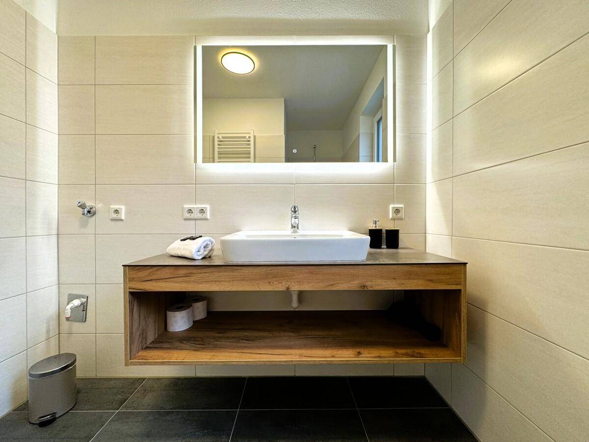 Modernes Badezimmer mit schwebendem Waschtisch, beleuchtetem Spiegel und hellen Fliesen.