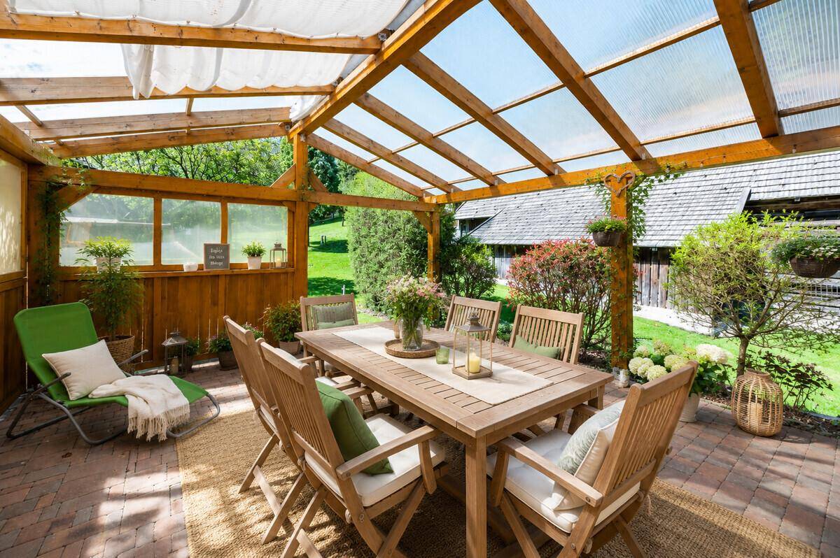 Gemütliche überdachte Terrasse mit Holztisch und Stühlen, umgeben von Grün.