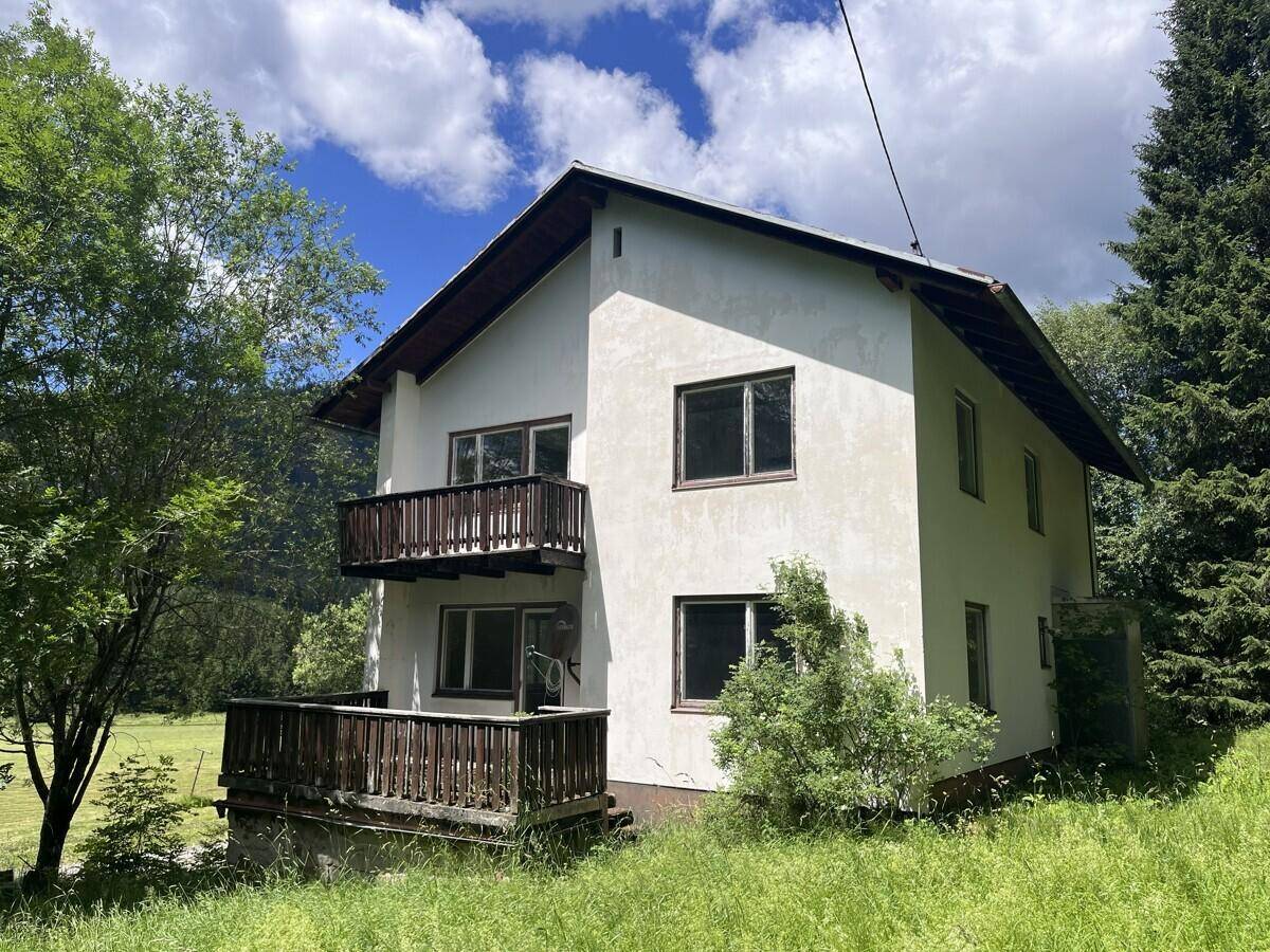 Haus mit Balkonen und ländlicher Umgebung unter blauem Himmel und grünen Bäumen.