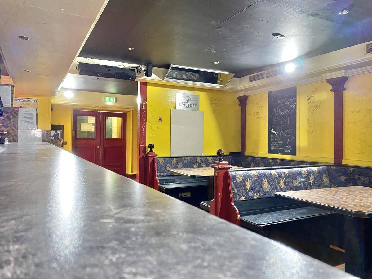 Innenansicht einer Bar mit gelben Wänden, roten Säulen, Sitzbänken und einer Theke im Vordergrund.