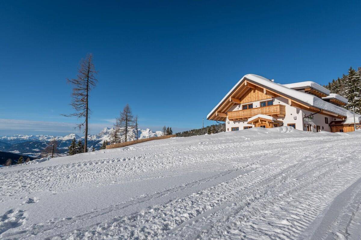 Alpenhaus in sonniger Winterlandschaft mit weitem Blick über verschneite Hänge und Berge.