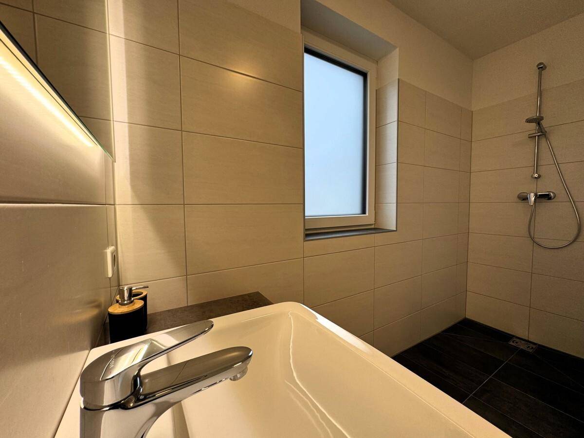 Modernes Badezimmer mit ebenerdiger Dusche, Fenster und hellen Wand- und Bodenfliesen.