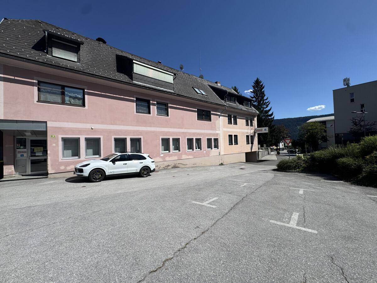 Vorderansicht des Gebäudes mit Parkplatz, einem weißen Auto und Blick auf die Berge.