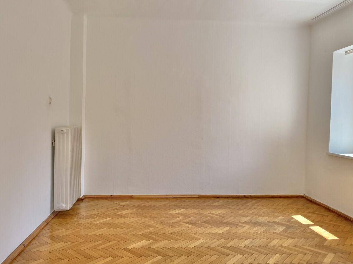 Geräumiges Zimmer mit hellem Fischgrätparkett und weiß gestrichenen Wänden, gut beleuchtet.