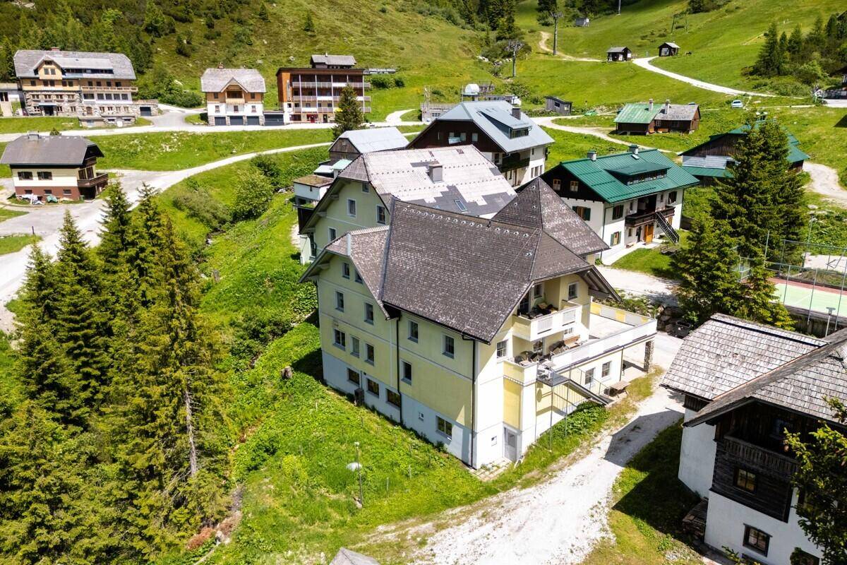 Luftaufnahme des gelben Gebäudes in einer grünen Berglandschaft mit weiteren Häusern und Bäumen.